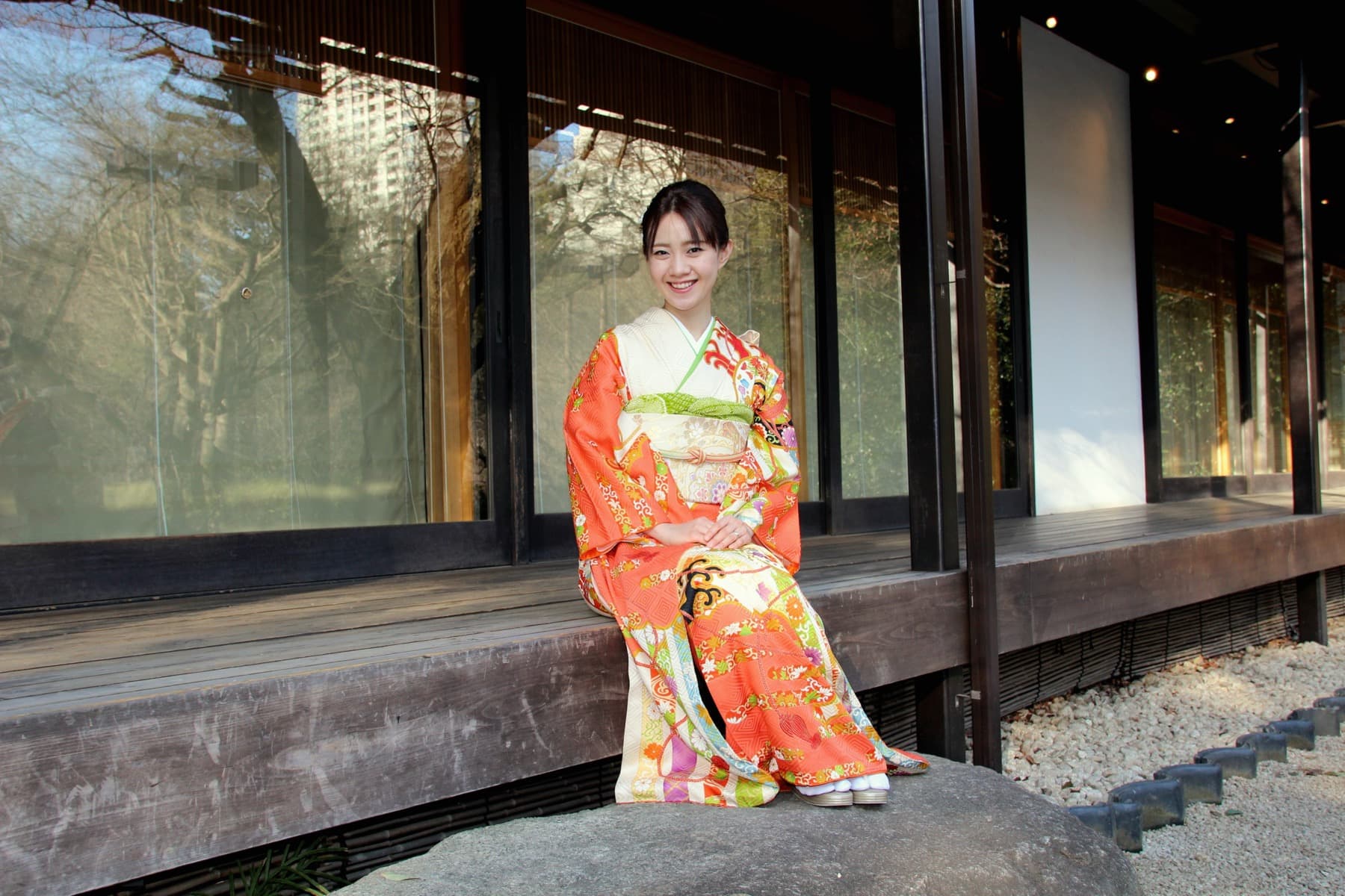 A woman in kimono sitting along an engawa veranda beside a traditional Japanese house.